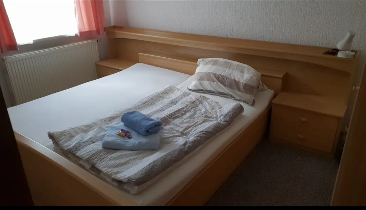BookNoFees: Ferienwohnung in Schönfeld, Sachsen, Deutschland – Schlafzimmer mit Holzbettgestell, gefalteten Handtüchern und einem Fenster mit rosa Vorhängen