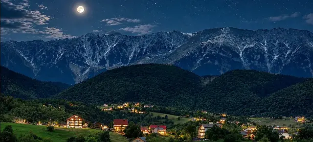 BookNoFees : location de vacances à Brasov – Vue nocturne d'un village de montagne avec ses maisons illuminées et la pleine lune